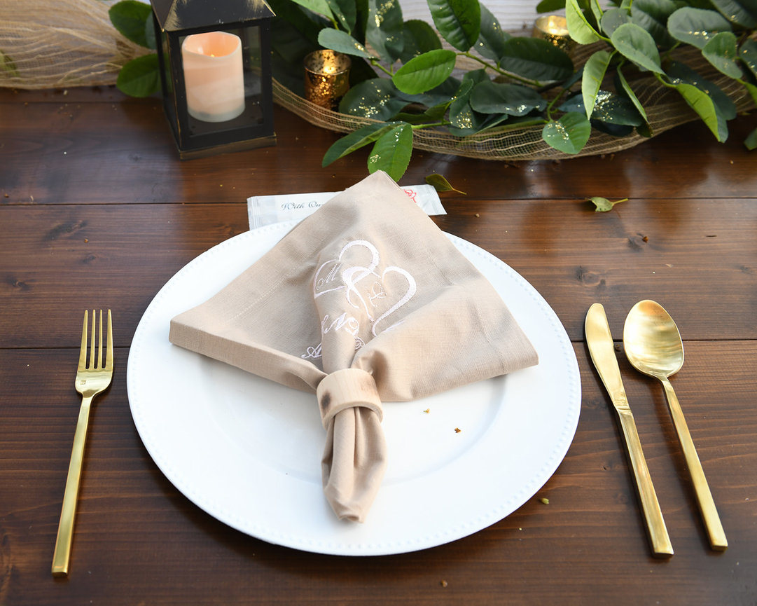Elegant table setting with a folded beige napkin, gold cutlery, and a lantern amidst greenery.