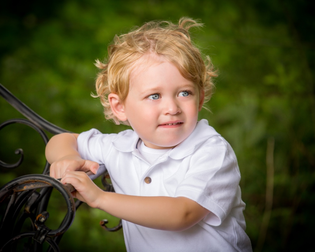 Young child with curly blond hair in a white shirt, holding a black metal railing outdoors with a green background.