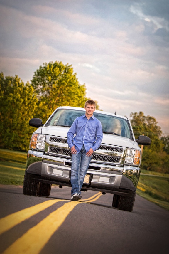 A person in a blue shirt leans against a white truck parked on a road with trees and a cloudy sky in the background.