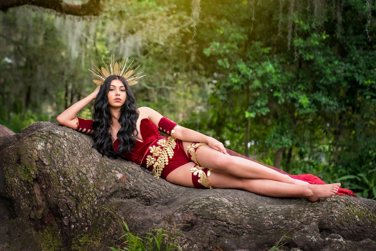 Woman in a red embellished gown poses on a tree trunk, wearing a golden crown in a lush outdoor setting.