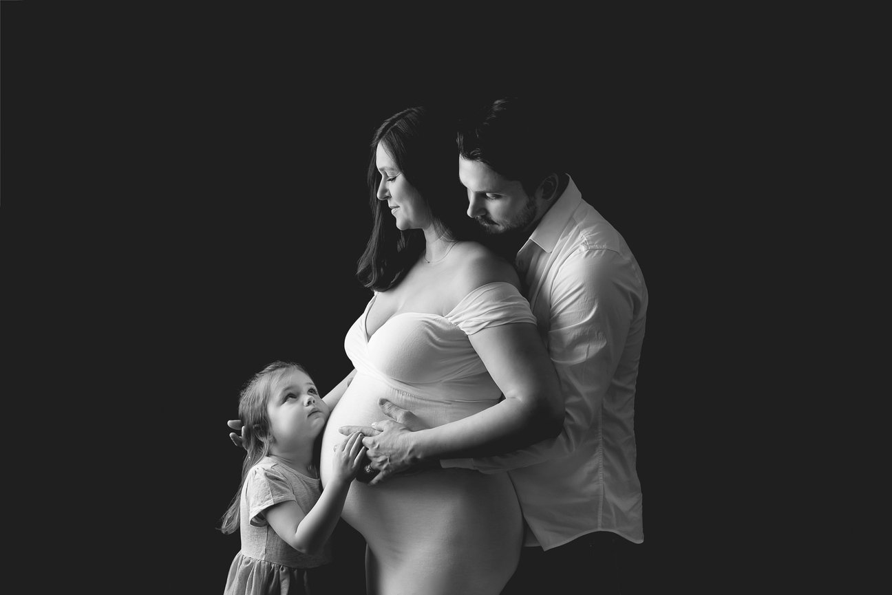 A black and white photo of a pregnant woman in a white gown, a man gently embracing her from behind, and a young child standing in front, touching her belly. The family appears calm and connected, set against a dark background.