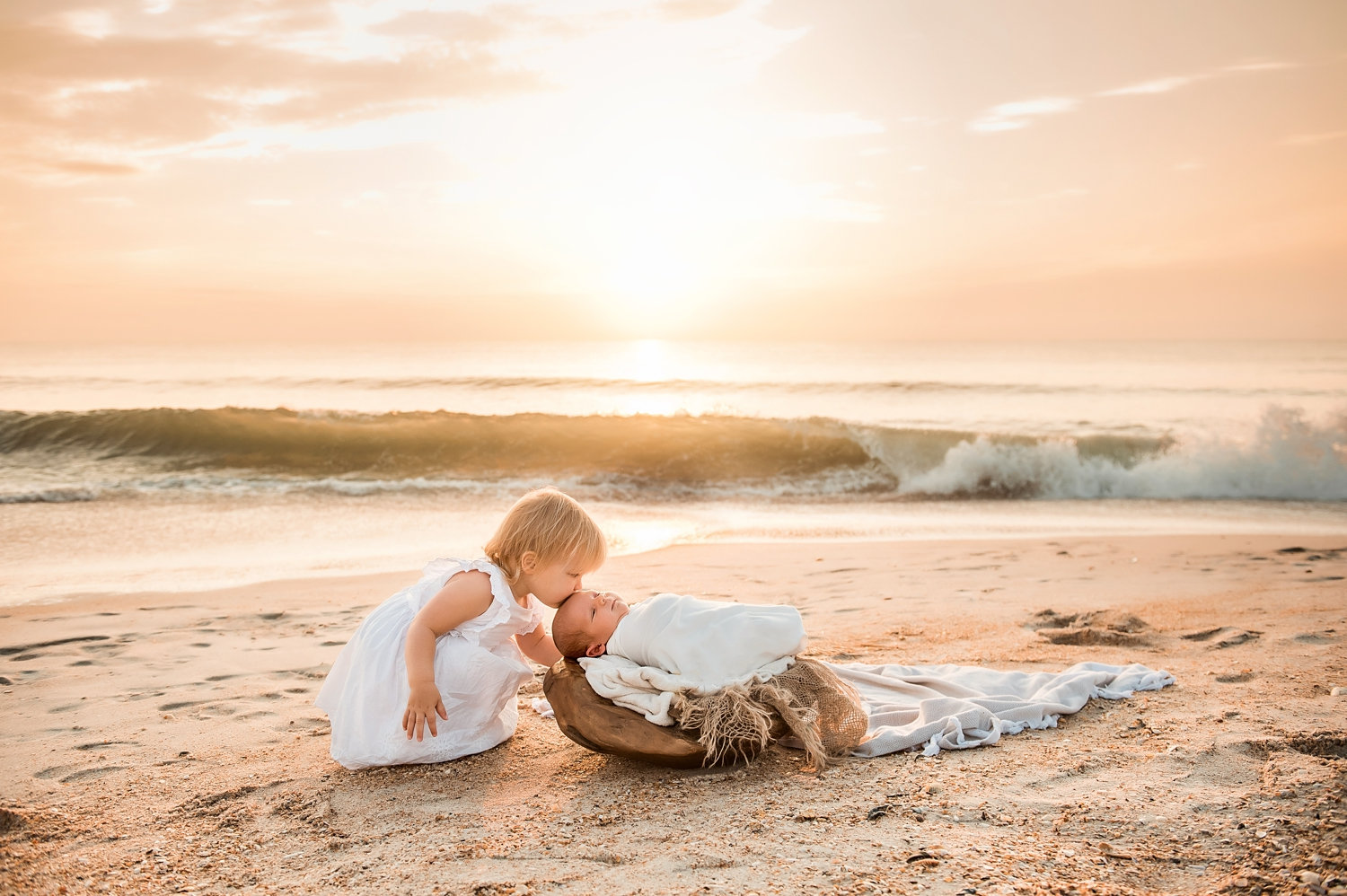 little girl kissing newborn baby sister on her forehead, the waves are rolling in the background, the sun is coming up over the Atlantic ocean