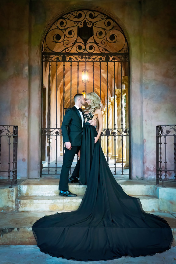 Elegant couple sharing a kiss in formal attire, with a dramatic black gown and ornate iron gates in the background.
