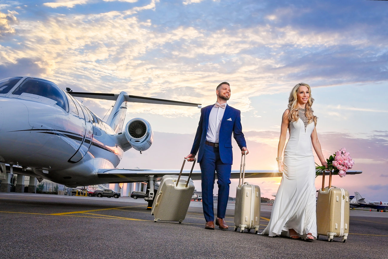 Couple in formal attire holding luggage and flowers, walking by a private jet at sunset.