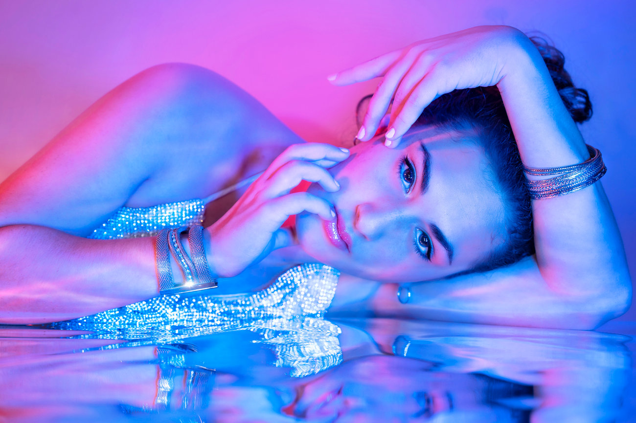 Woman in a sparkly outfit poses gracefully with a hand on her face, surrounded by colorful blue and pink lighting.