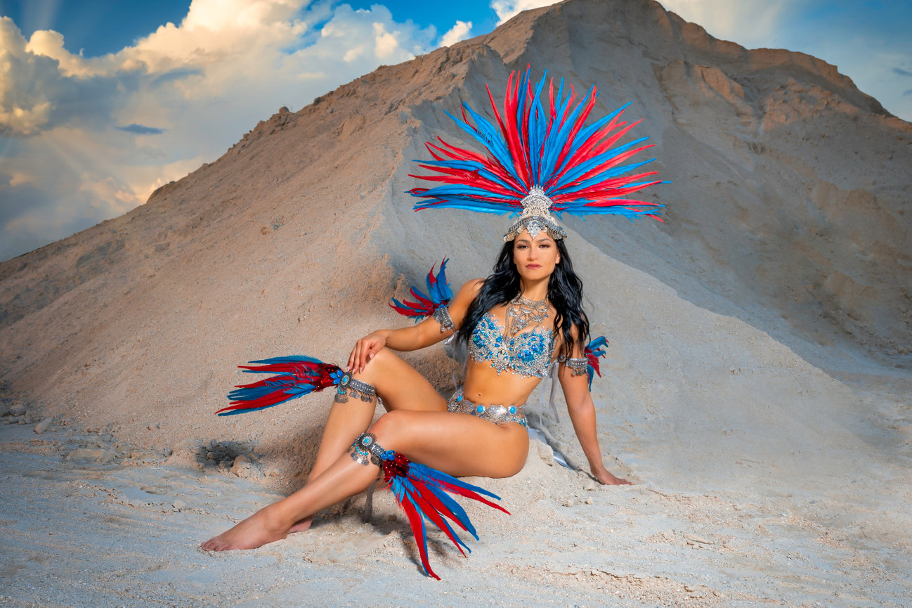 Person in vibrant costume with feathers posing on sand, against a backdrop of hills and clouds.
