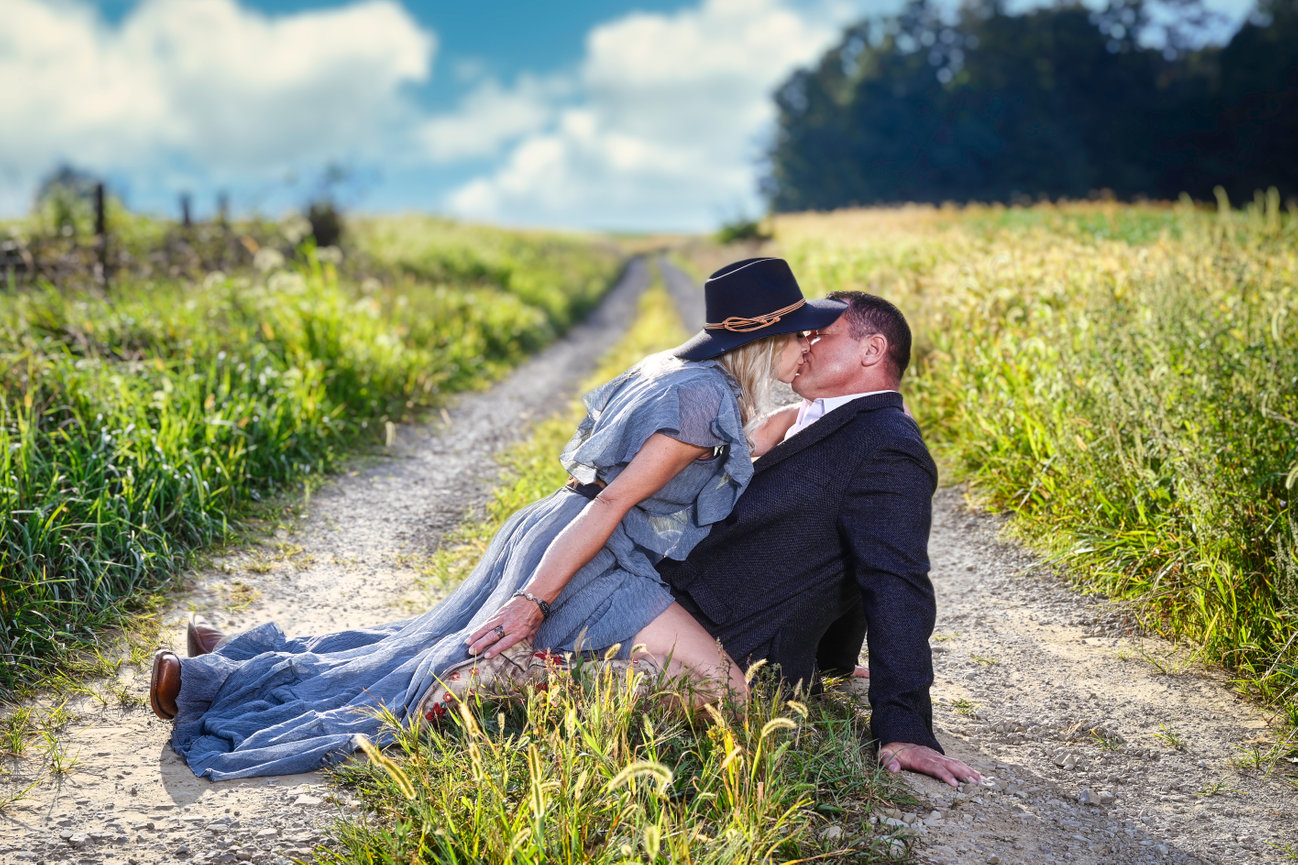 A couple embraces on a dirt road surrounded by tall grass, set against a blue sky and trees.