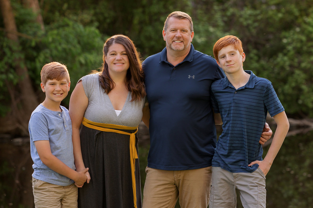 Family of four posing outdoors, with trees in the background, smiling at the camera.