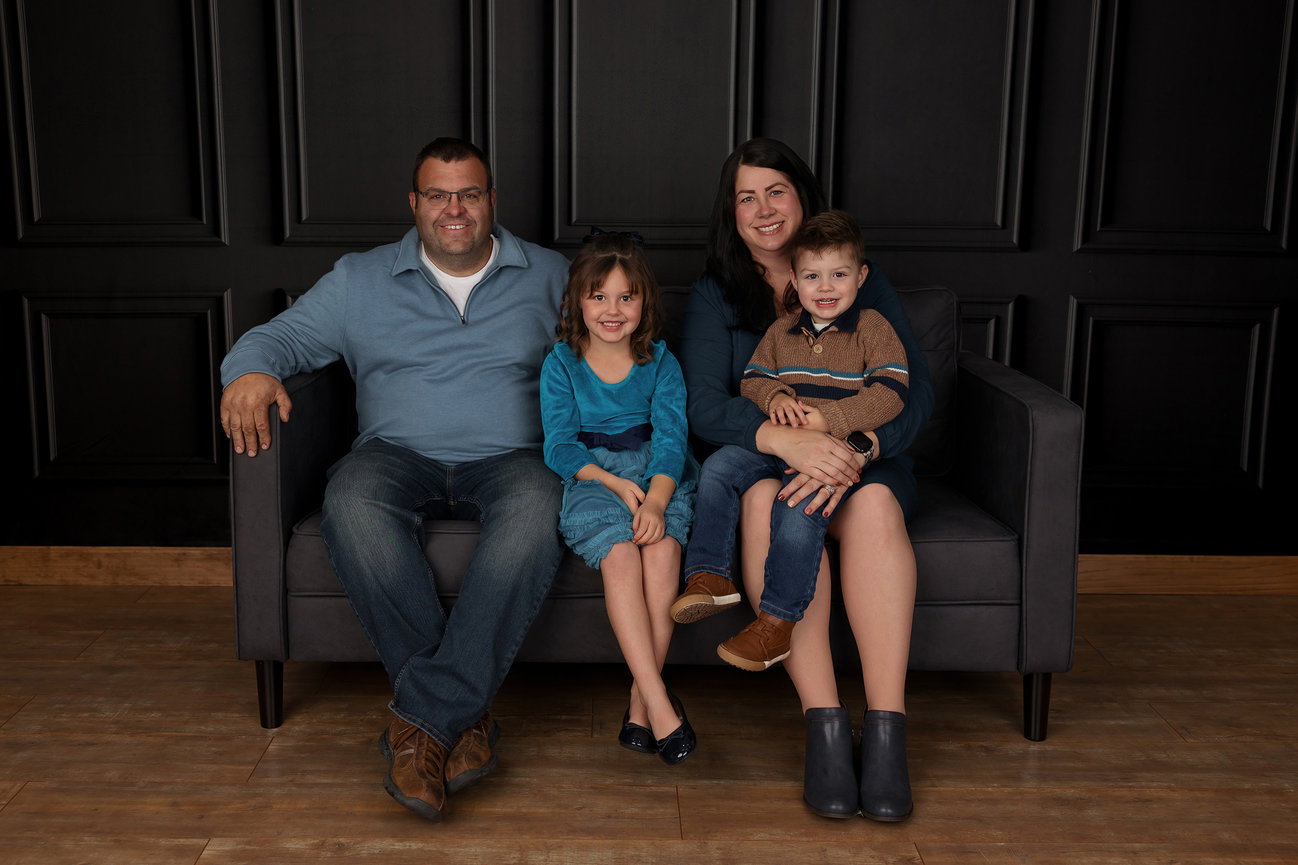 Family of four sitting on a gray sofa against a dark paneled wall.