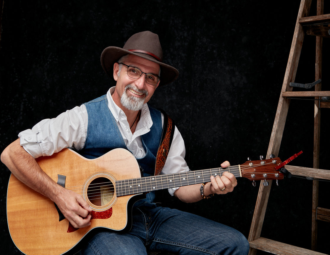 Man in a hat playing an acoustic guitar with a wooden ladder in the background.