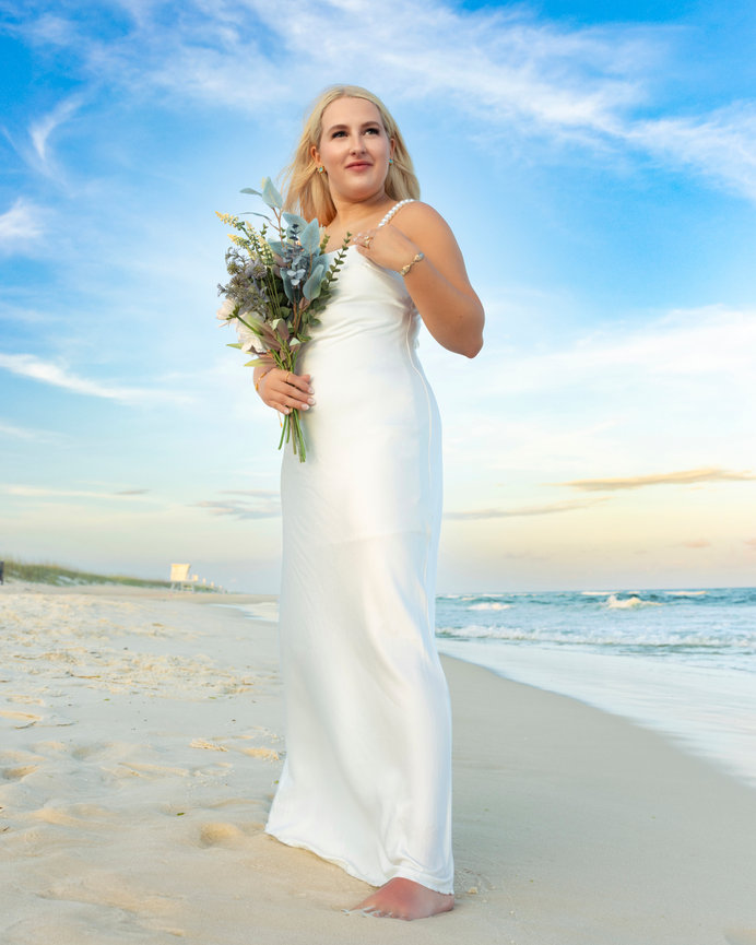 Bride walking barefoot on the beach in a satin gown, holding a bouquet during a Gulf Coast elopement photographed by Dixon Creative Images.