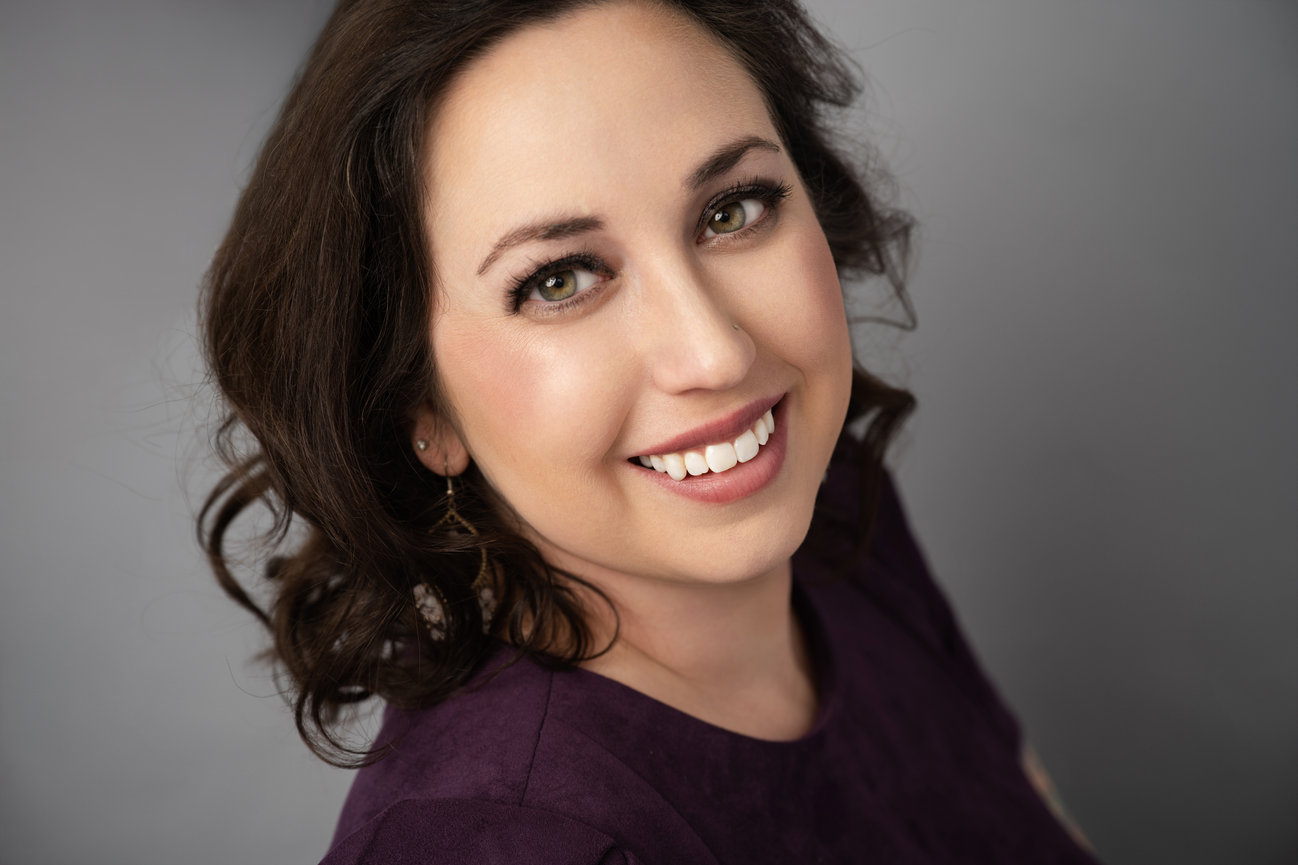 Smiling woman with wavy hair wearing a purple top against a gray background.