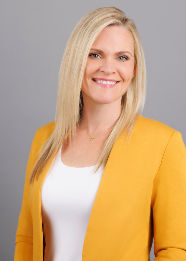 A person smiling, wearing a yellow jacket and a white top, with long blonde hair against a gray background.