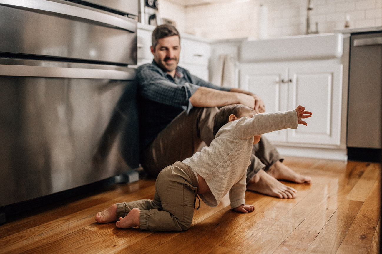 A family smiles at his crawling baby on a hardwood floor in a kitchen during a Huntsville Alabama family photoshoot.