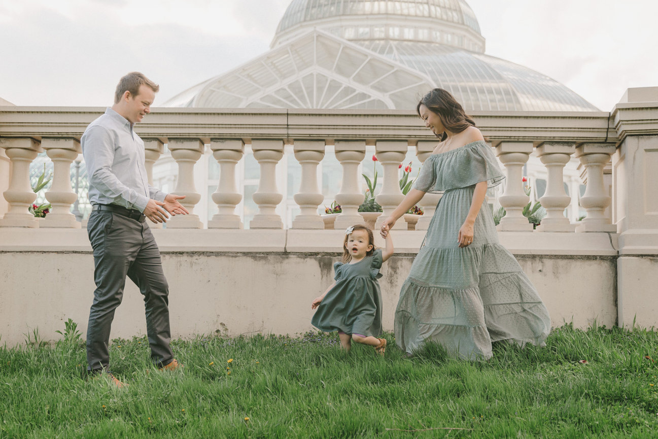 Family playing on grass near a conservatory; parents and toddler enjoy a joyful moment together.
