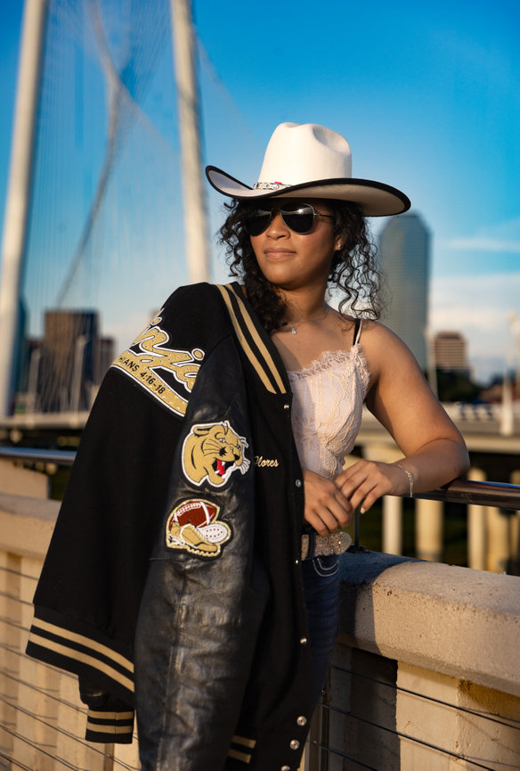 High School Senior Girl Standing on bridge wearing sunglasses and a cowboy hat with letterman jacket draped over shoulder