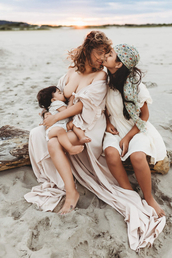 A mother sitting on driftwood breastfeeding her baby on the beach in Sandy Hook, New Jersey.