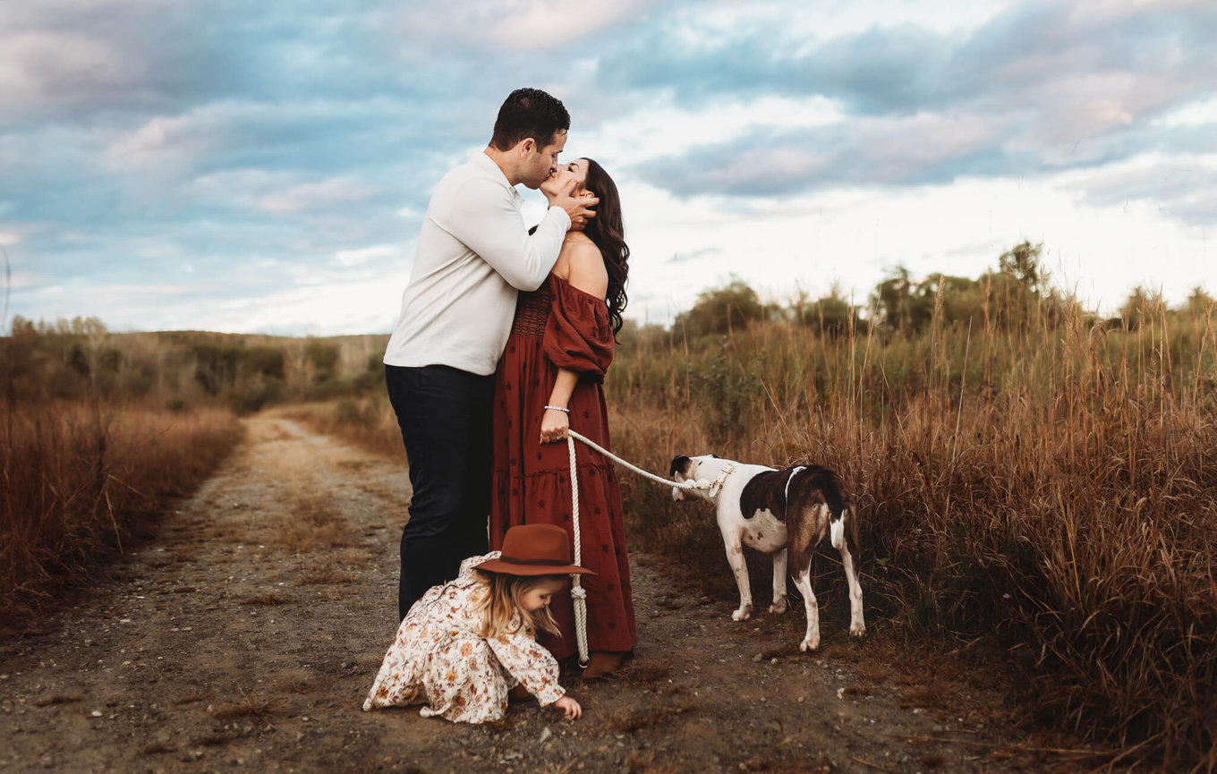 A husband and wife kissing in a field under a blue sky while their daughter and dog play around them in Morris County, New Jersey.