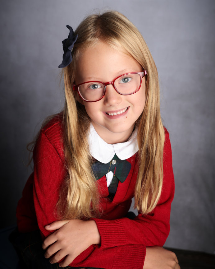 A smiling child with long blonde hair, red glasses, and a red sweater, seated against a neutral background.