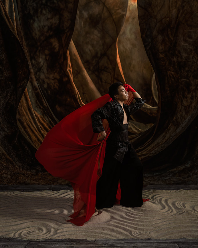 Person in dark clothing with a flowing red cape, standing on a textured surface in front of a dramatic, draped backdrop.