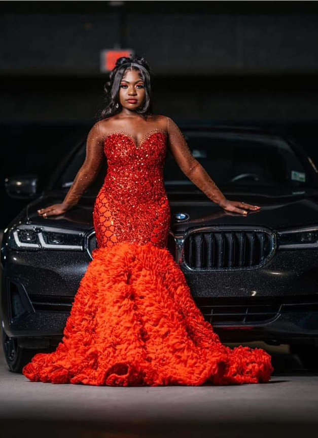 Woman in a red gown stands confidently in front of a black luxury car.