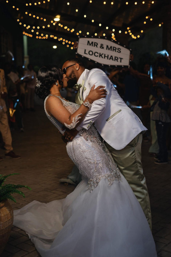 A couple in elegant wedding attire shares a kiss under string lights, surrounded by people, with a Mr & Mrs sign in the background.