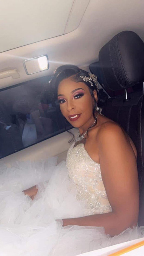 Woman in a strapless white wedding dress sits in a car, smiling and looking at the camera.