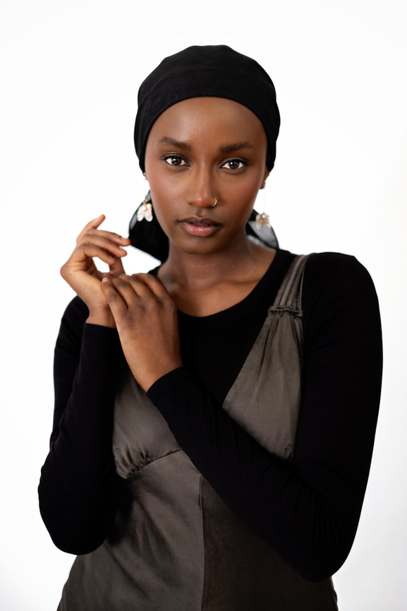 A person wearing a black headscarf, black top, and brown dress with a white background.