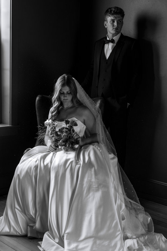 Bride sitting in a chair holding a bouquet, with a groom standing beside her in formal attire, captured in black and white.