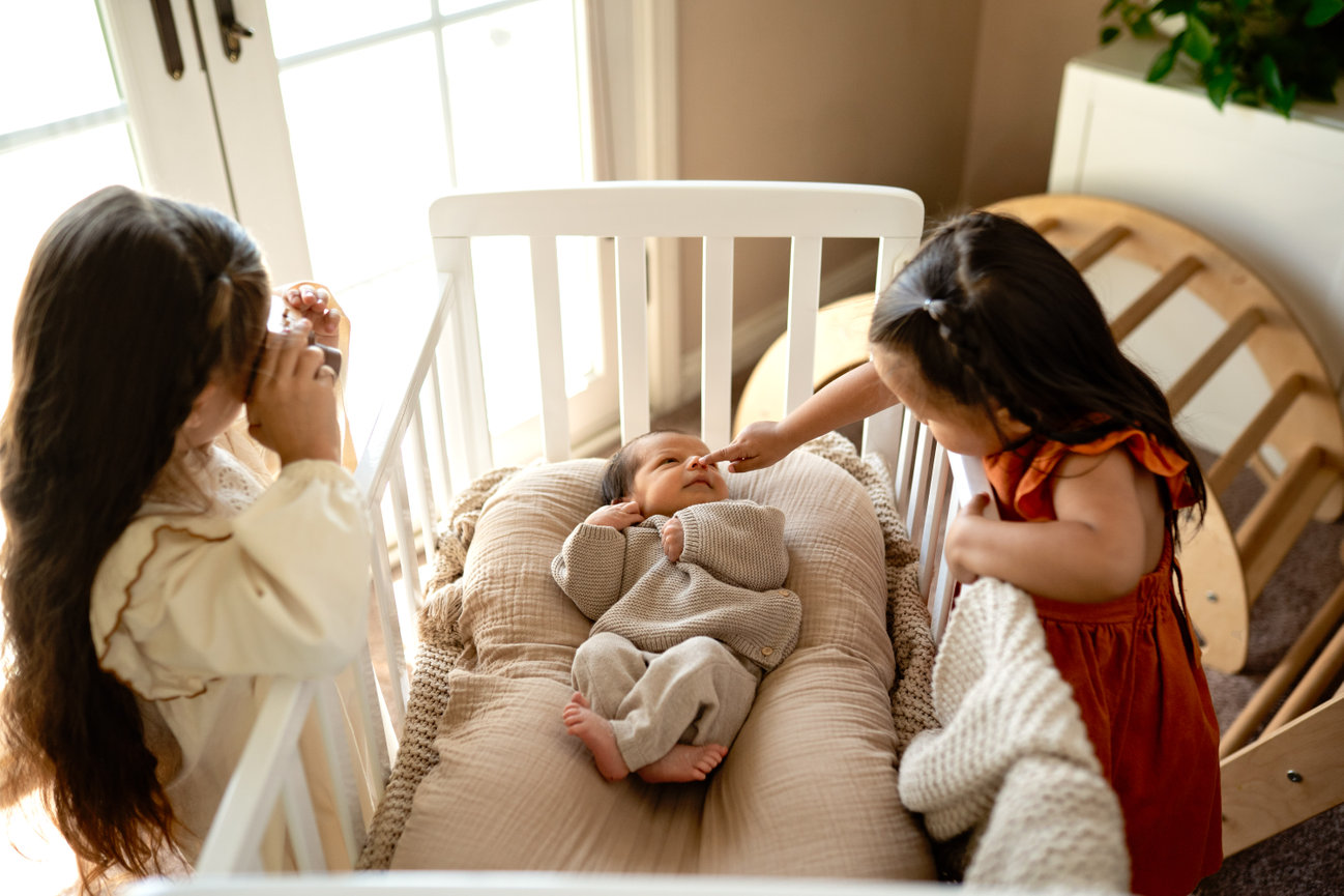 Two young girls looking at a baby lying in a crib by a sunlit window