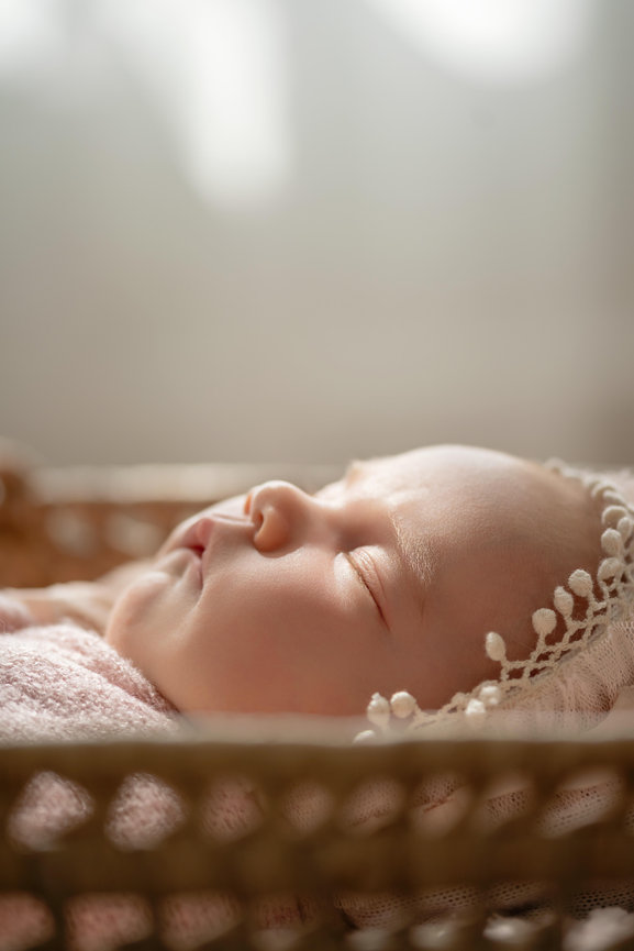 Sleeping newborn baby in a wicker basket with soft lace bonnet and warm sunlight.- newborn session San Diego