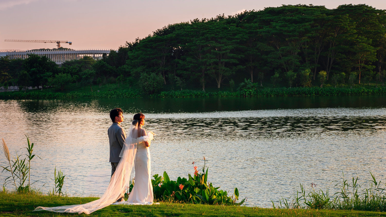 Bride and groom stand together beside a lake at sunset.