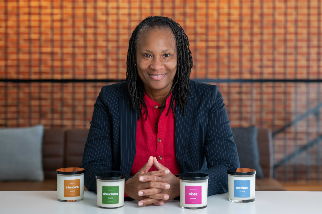 Smiling woman in blazer seated behind four labeled candles on a table.