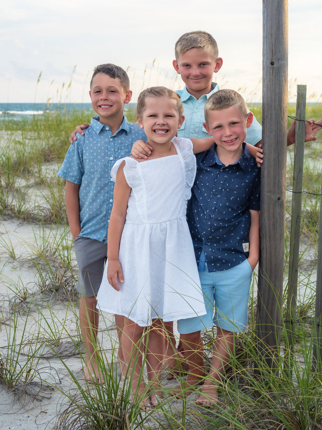 Four smiling child cousins stand together next to a fence in the dunes with an ocean background.