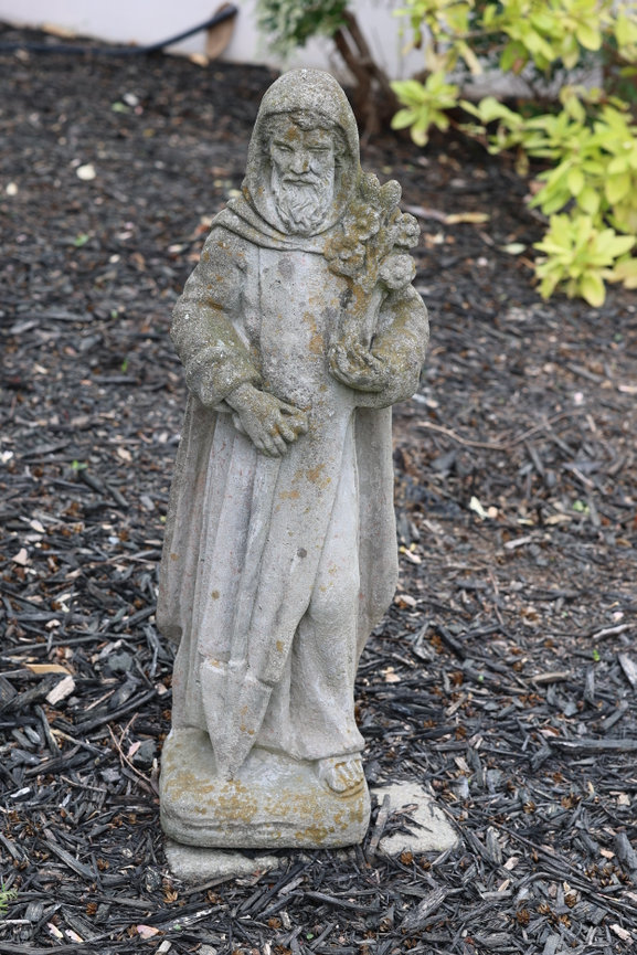A small stone statue stands beside plants in a garden bed.