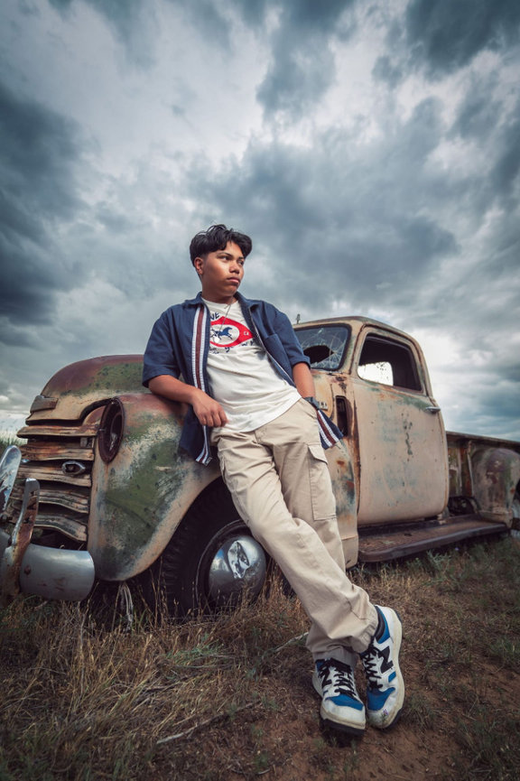Man leaning on a rusty vintage truck in a field under a cloudy sky.