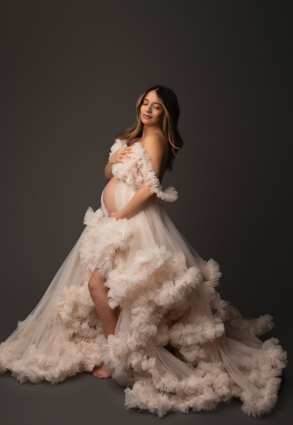 Pregnant woman in a flowing, ruffled gown poses gracefully against a dark background.
