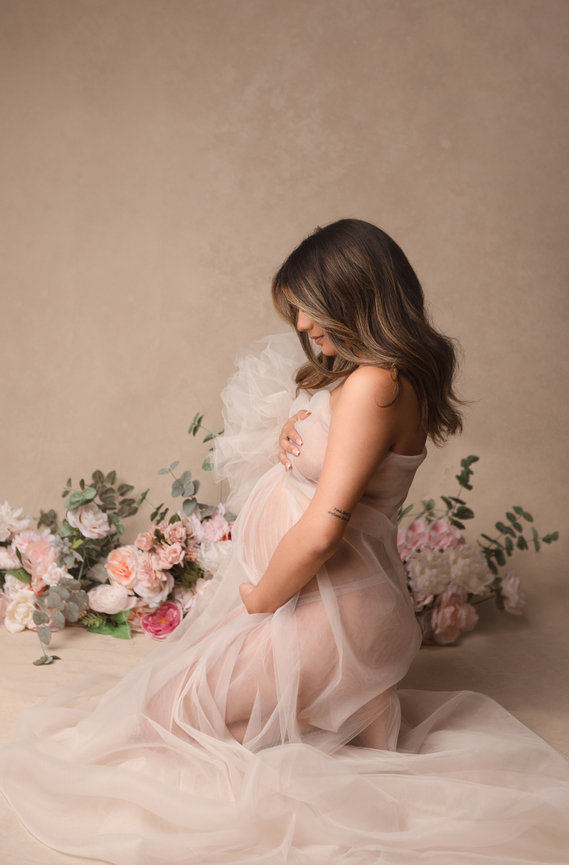 Pregnant woman kneeling in a sheer dress surrounded by pastel flowers against a neutral background.