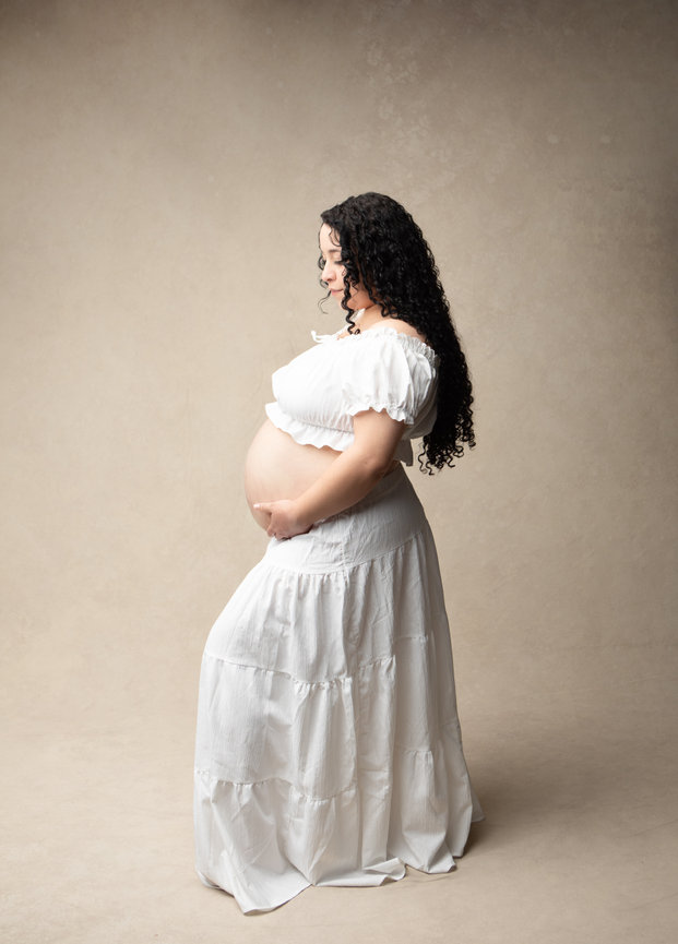 Pregnant woman in a white dress posing sideways against a neutral background.