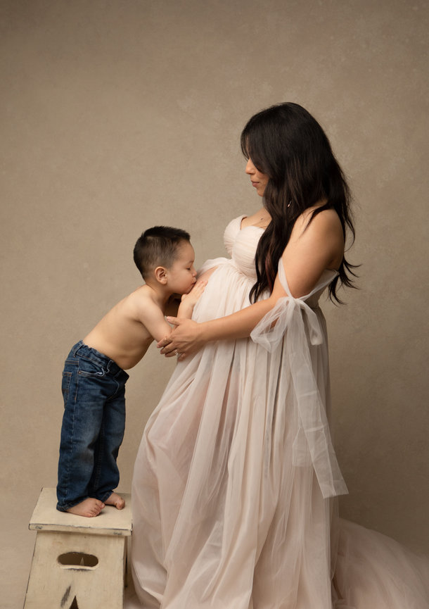 A pregnant woman in a flowing dress stands by a young boy kissing her belly.