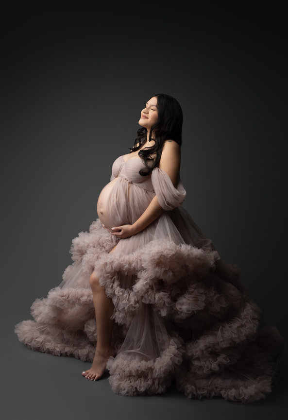Pregnant woman in a flowing, layered tulle gown poses with a serene expression against a dark background.