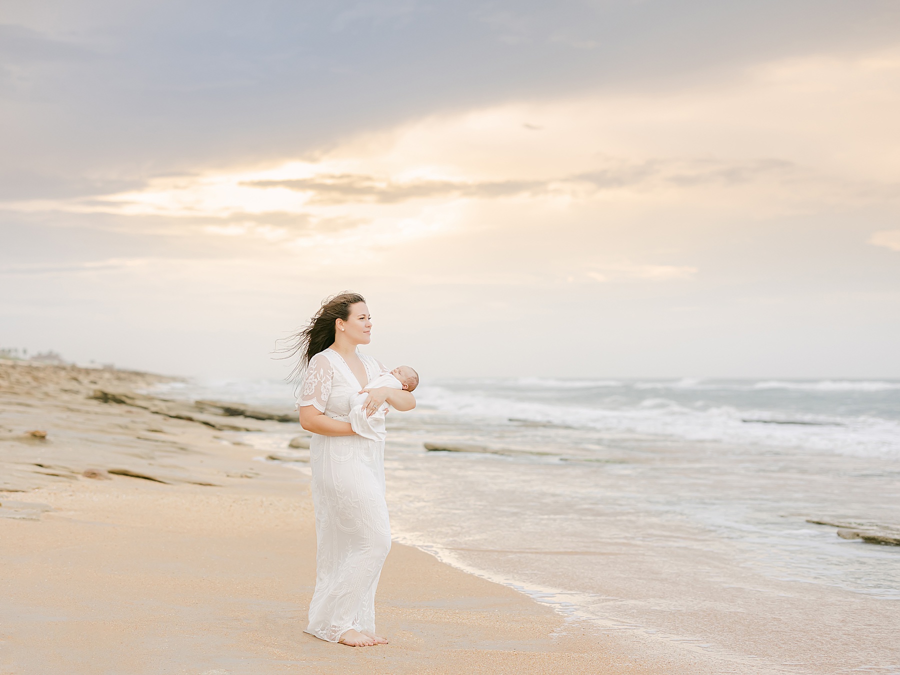 woman standing on the sand in a white dress holding a newborn baby