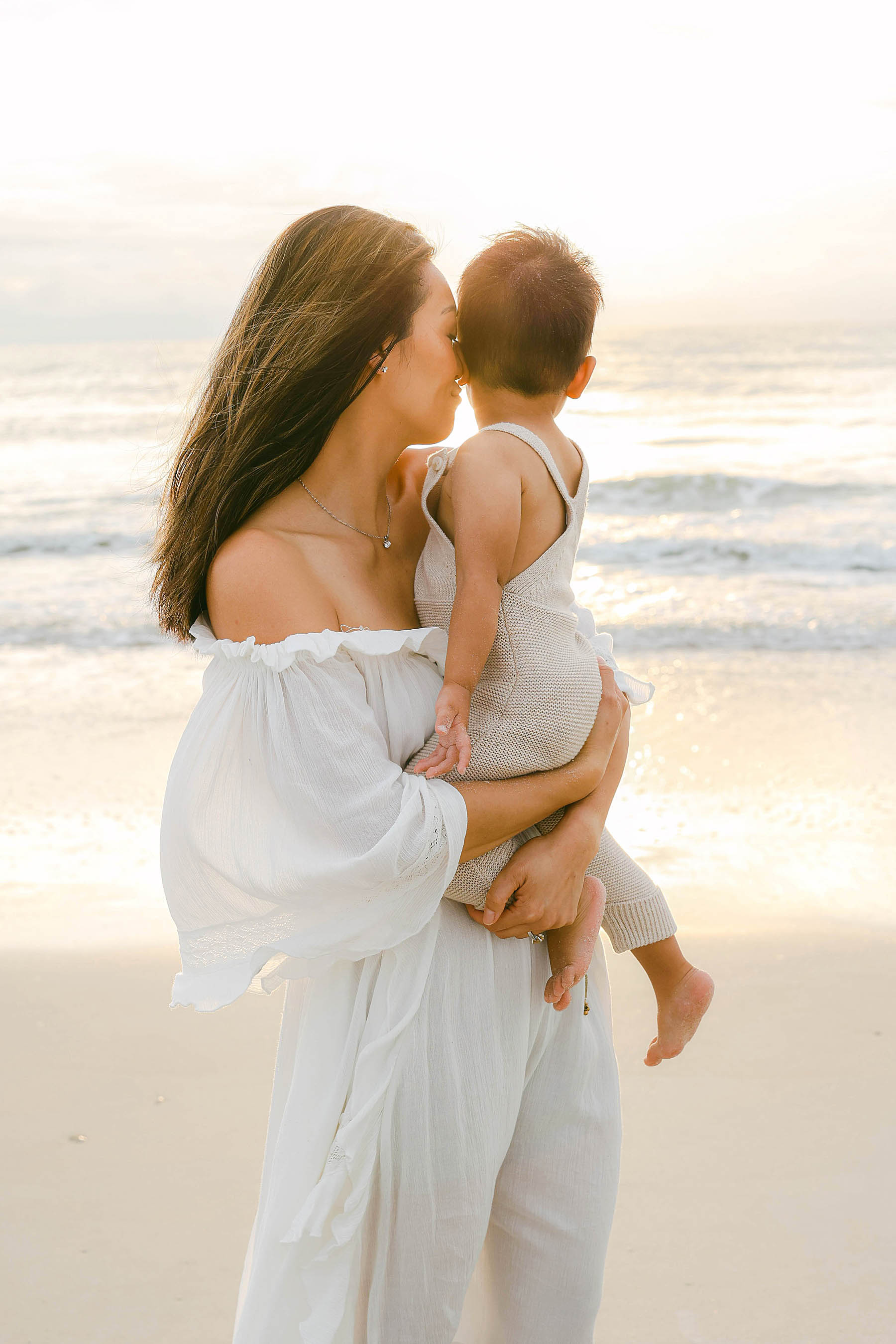 mom holding baby boy wearing white dress at sunrise on the beach