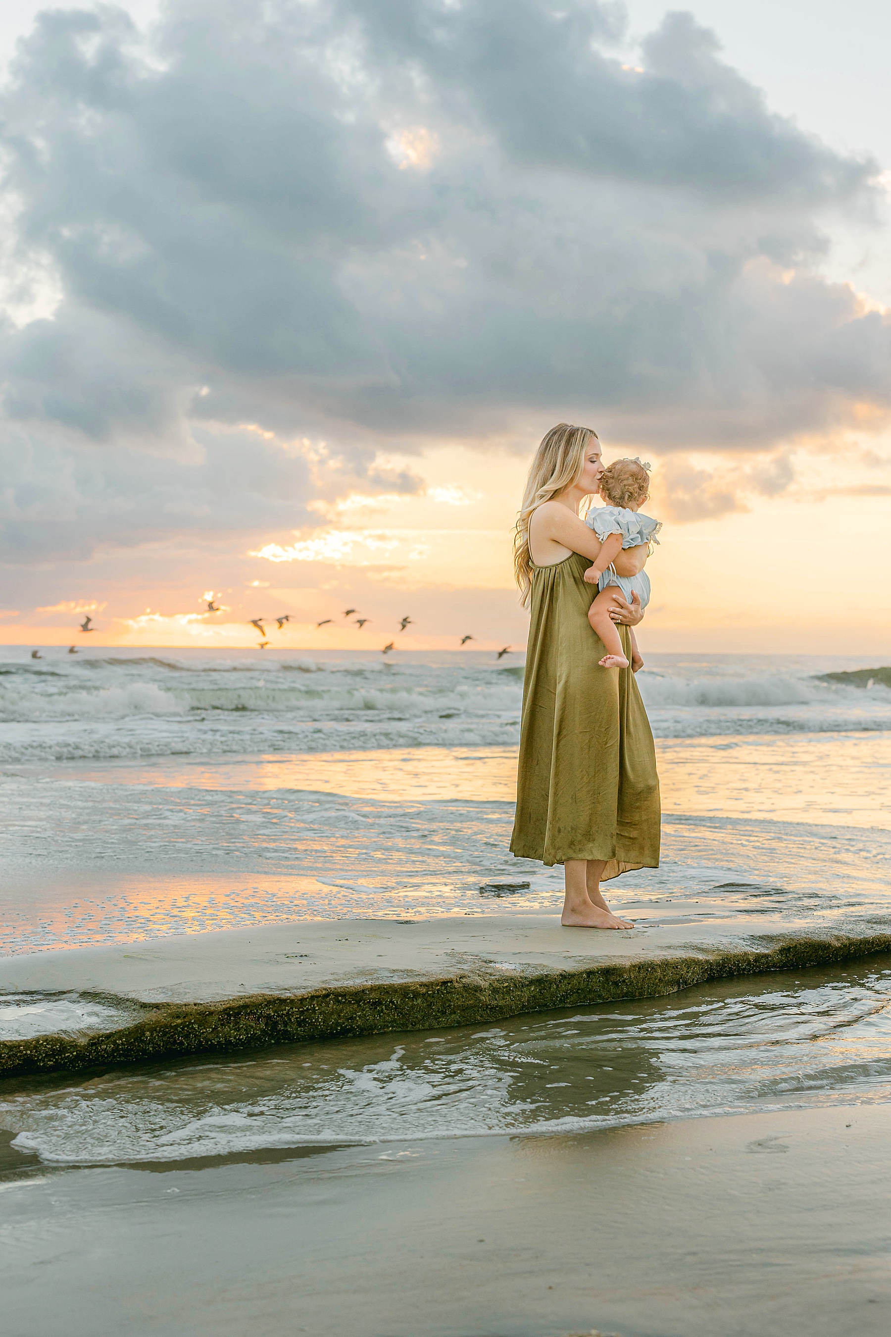 woman holding baby at the beach at sunrise wearing green dress