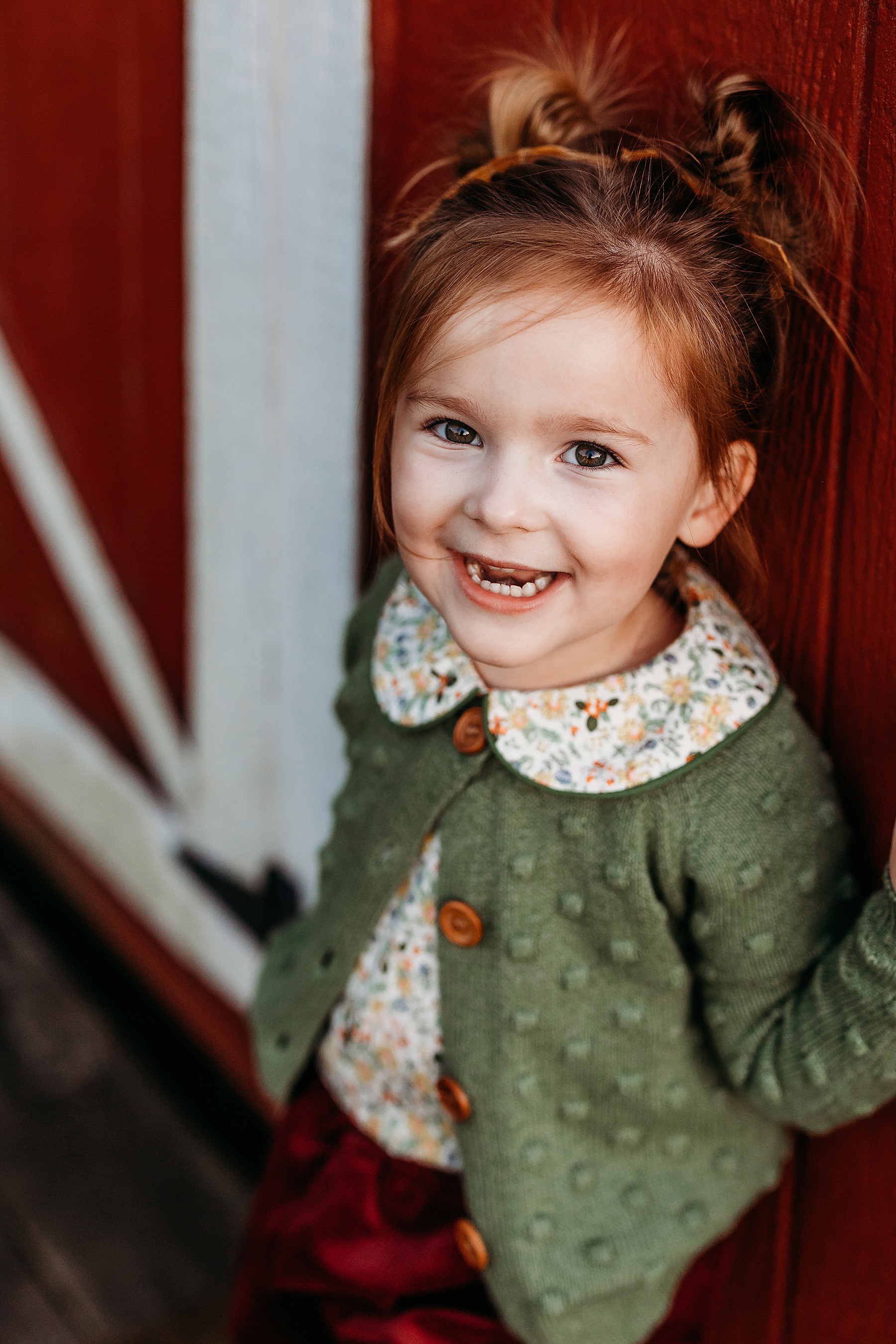 little girl in green sweater standing against a red barn door smiling