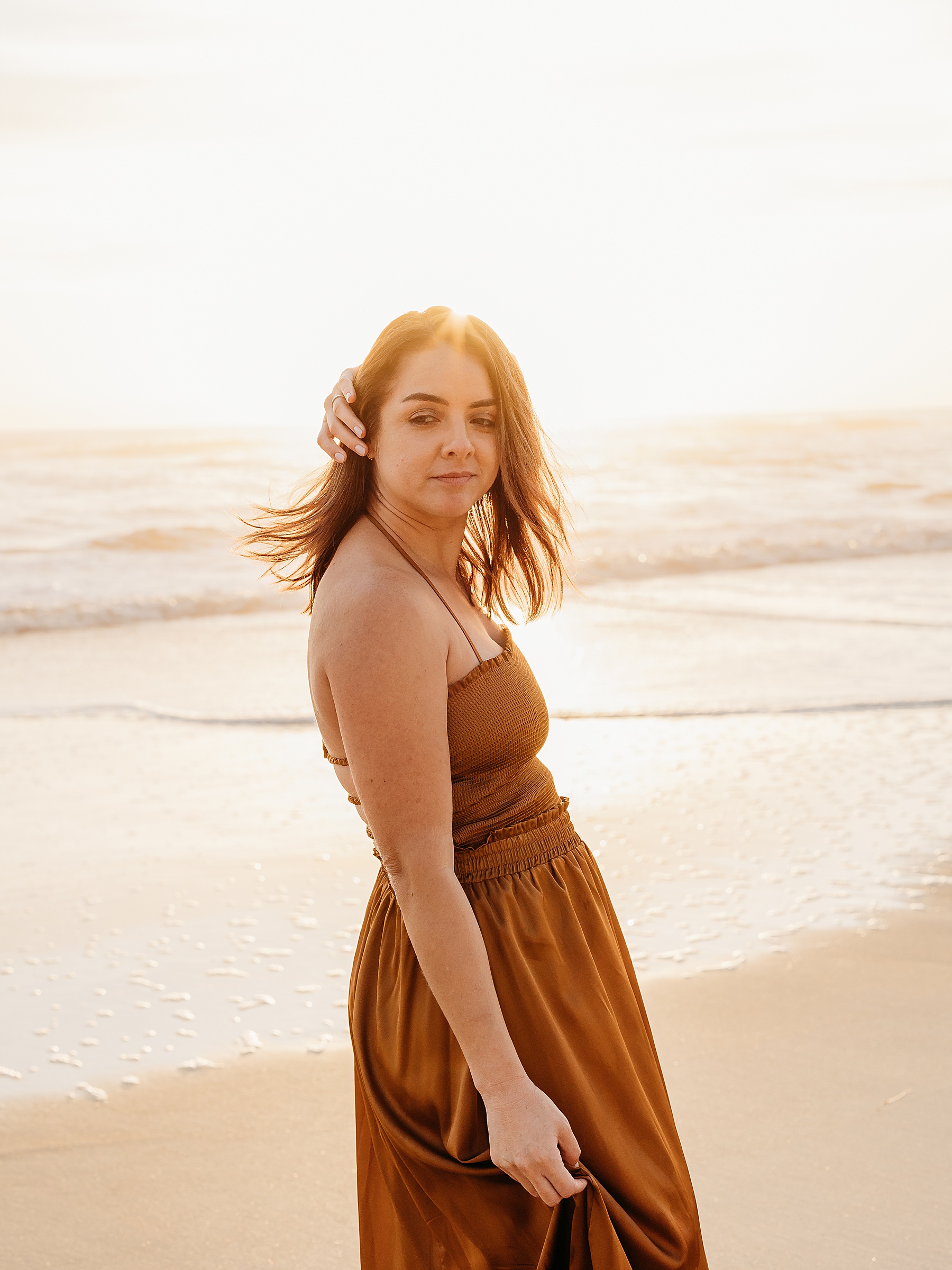 woman wearing rust colored dress on the beach at sunrise holding her hair