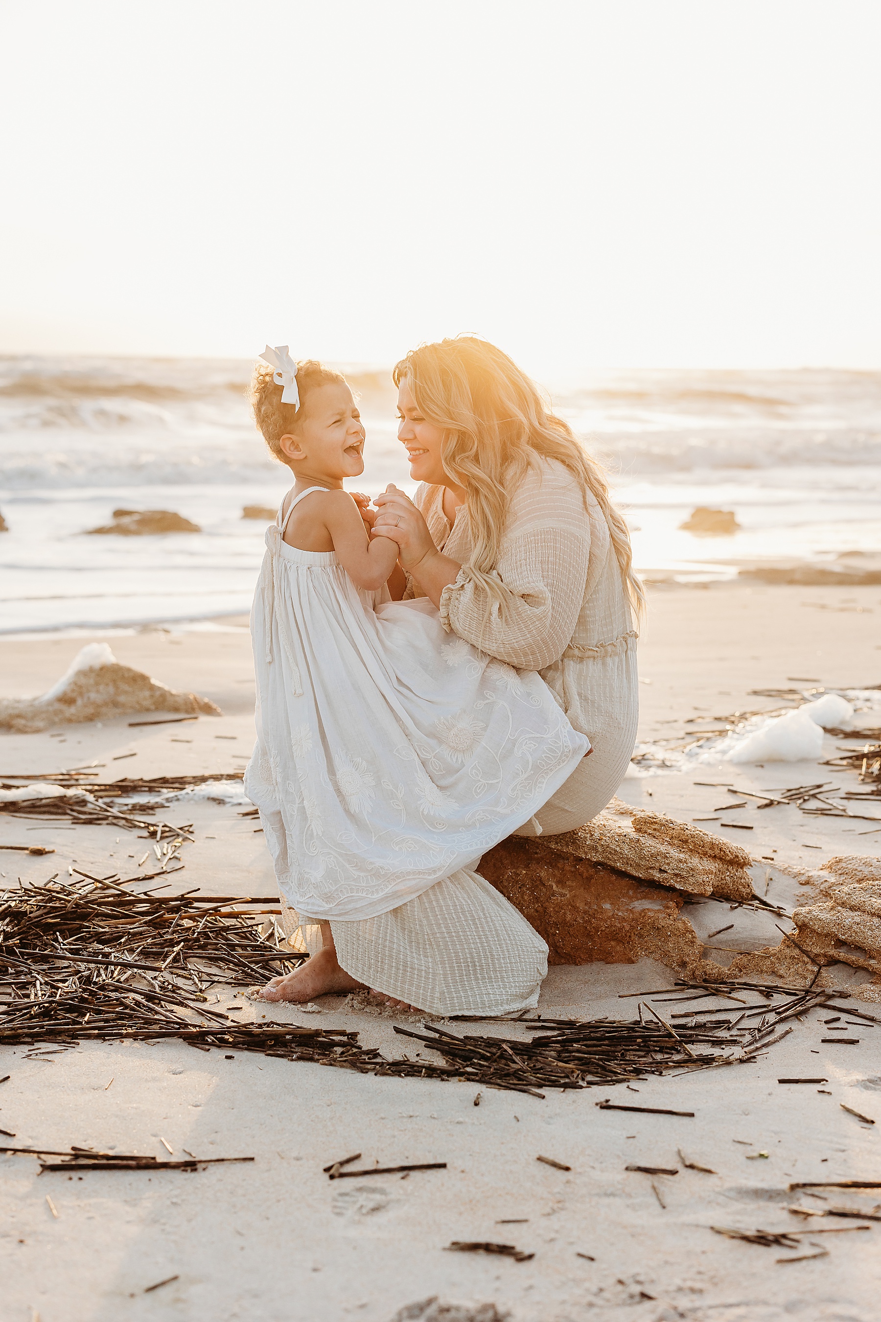 woman dressed in neutral long dress holding little girl on rock at beach at sunrise