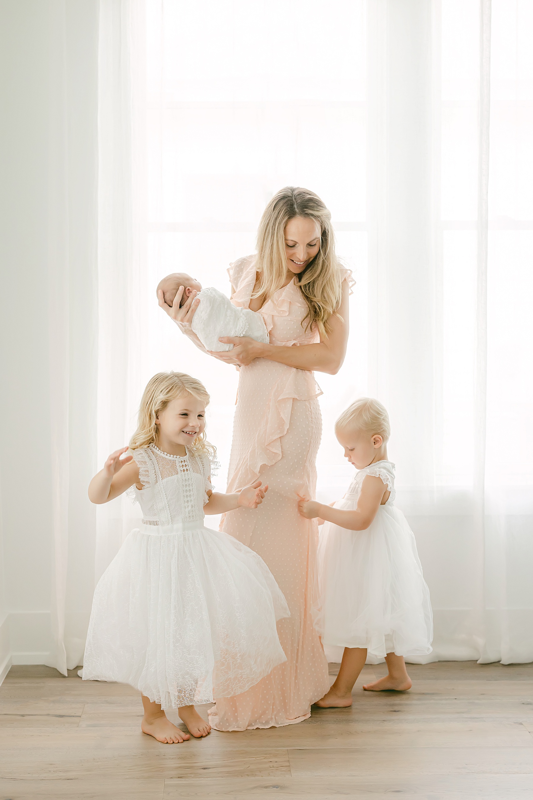 woman holding a newborn baby girl standing in front of window with sheer curtains while little girls dressed in white dance around her