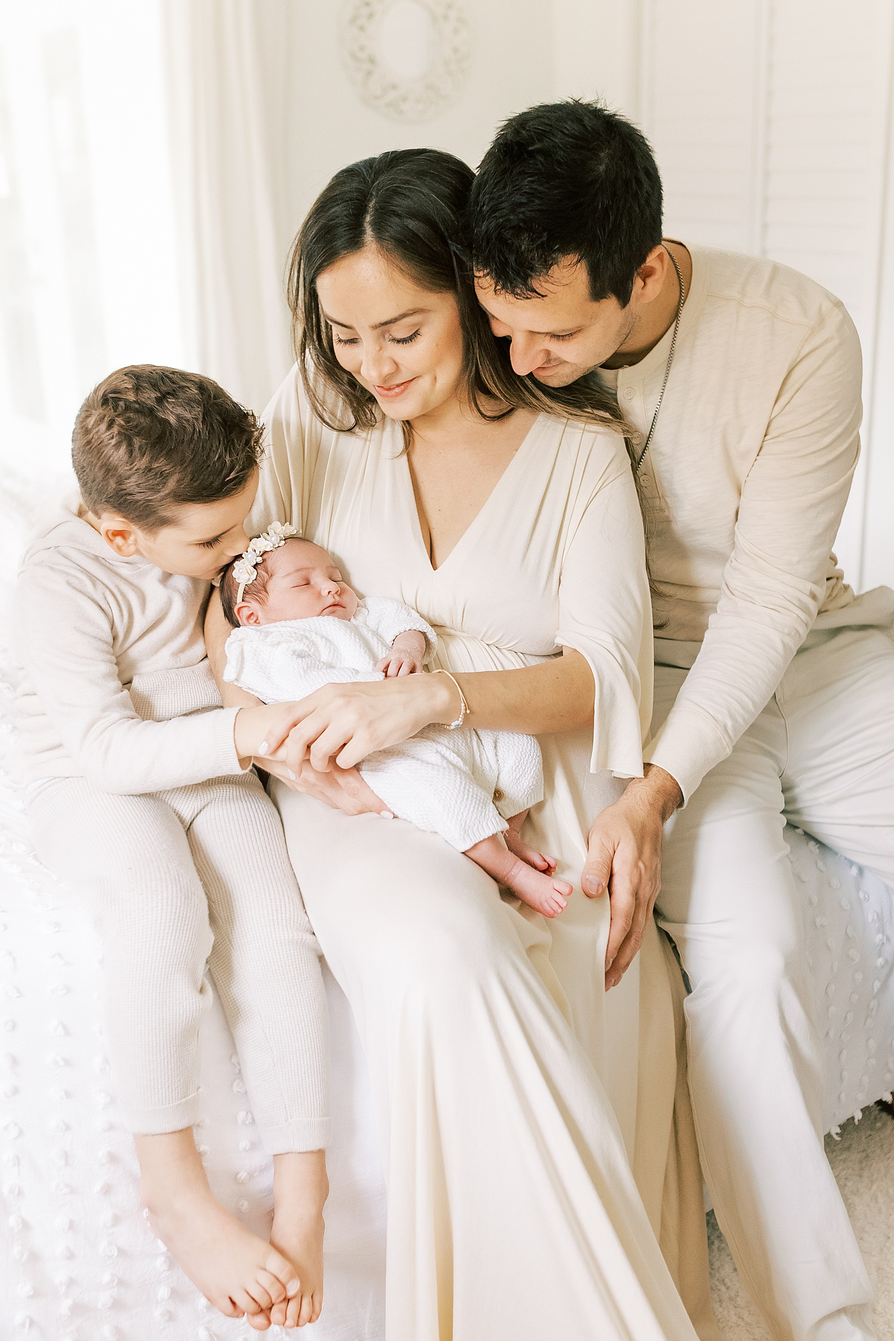 family wearing neutral colors sitting on white bed holding newborn baby girl