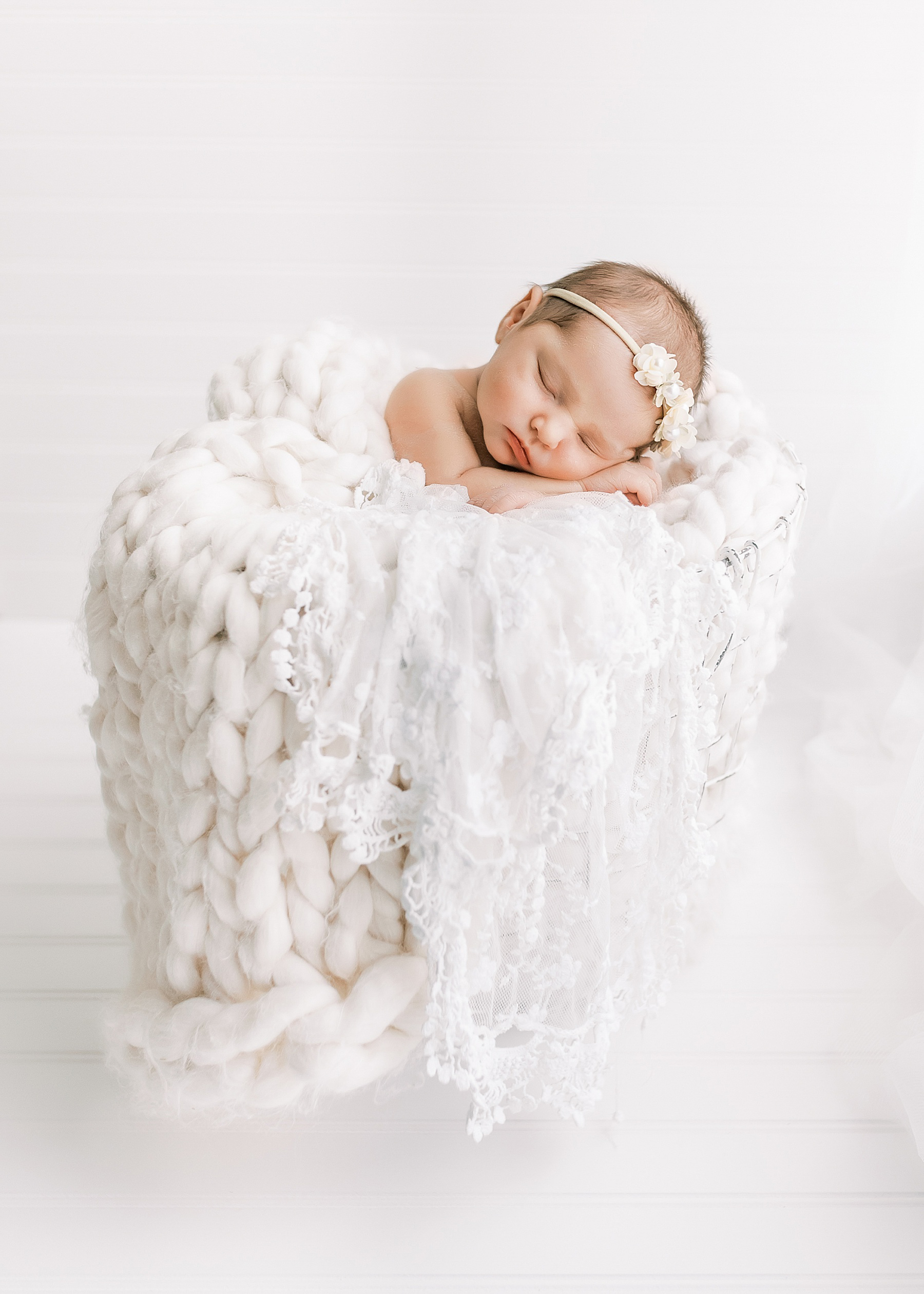 newborn baby girl wrapped in white blankets in white wire basket on white floor in front of a window